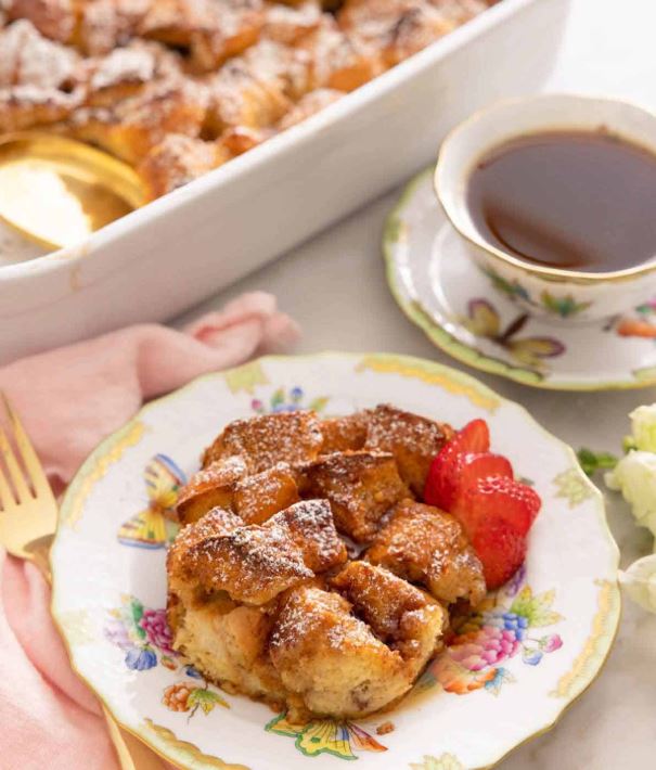 A plate of French toast casserole alongside a cup of tea and a casserole dish filled with French toast casserole. 