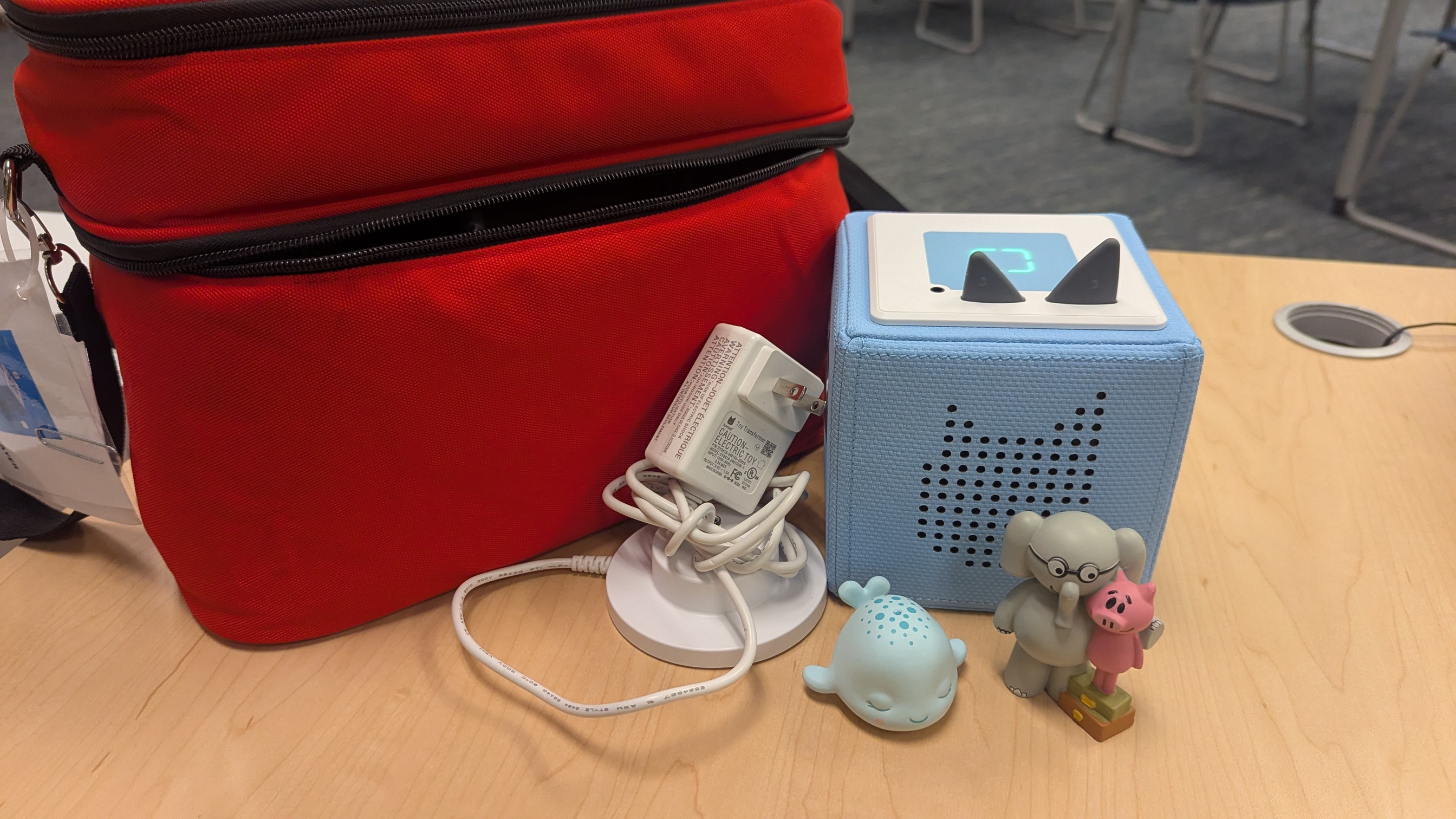 Displays a red carrying case, Toniebox, Sleepy Whale, and Elephant & Piggie figurine along with a charging cable 