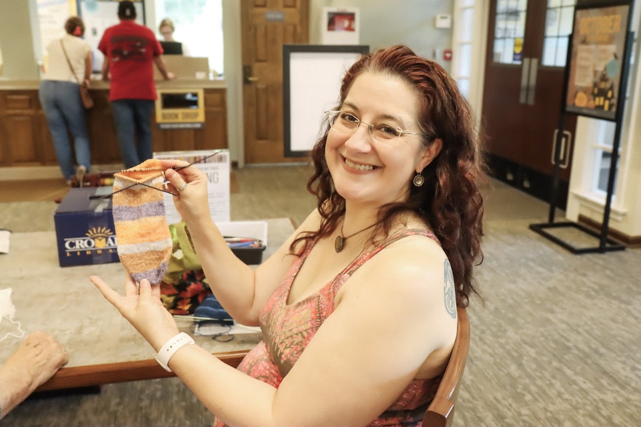 A community member holding up a sock she is knitting in the Library. 