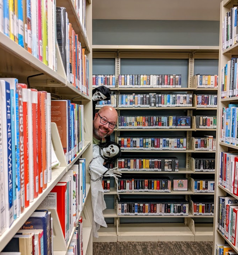 Librarian Erik poking out from behind a bookshelf with two ghosts. 
