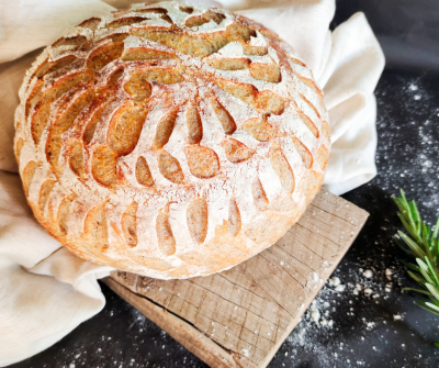 A loaf of sourdough bread on a small wooden cutting board, next to a sprig of rosemary. 