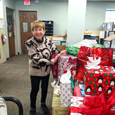 Mary Lynn standing in front of a huge pile of gifts, donated as a part of the Giving Tree project in 2024. 