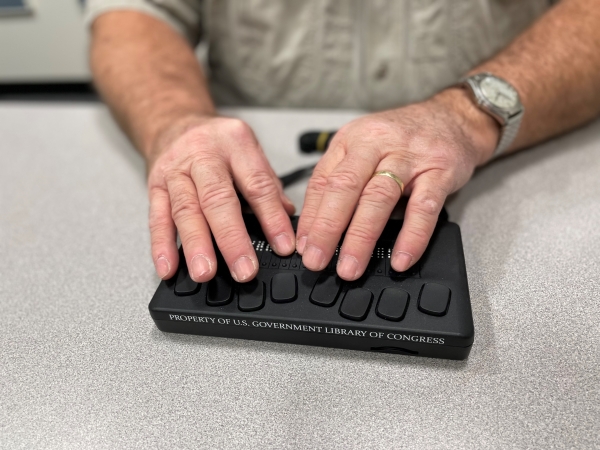 Two hands sit atop a Braille tool, part of the Braille and Talking Book Library Service. 