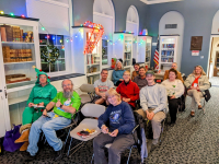Happy folks sitting in rows of chairs during an Ale Together Now session, in the decorated Community Room.