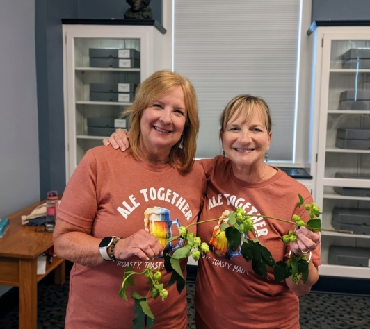 Mary and Terry, patrons of Ale Together Now, wearing matching "Ale Together Now" t-shirts and holding a vine of hops. 