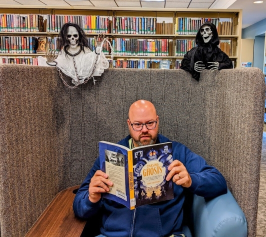 Librarian Erik reading a spooky ghost book, completely unaware that there are two ghosts hovering over his chair. 