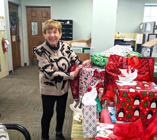 Mary Lynn standing in front of a huge pile of gifts, donated as a part of the Giving Tree project in 2024. 