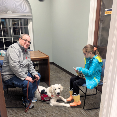 A girl in a light blue coat reads aloud from a book to a white service dog laying at her feet. The dog's handler sits in a chair across from her. 