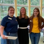 A teen stands in the Youth Program Room between Librarians Lucy and Kelsey, receiving her scholarship certificate. 