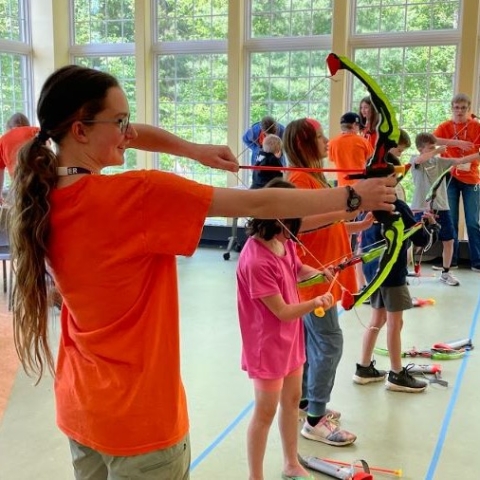 A teen volunteer shooting a toy arrow, along with a group of kids, during Percy Jackson Camp in the Youth Program Room. 