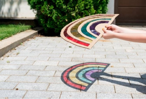A child's hand holding a handmade rainbow suncatcher over the pavement. 