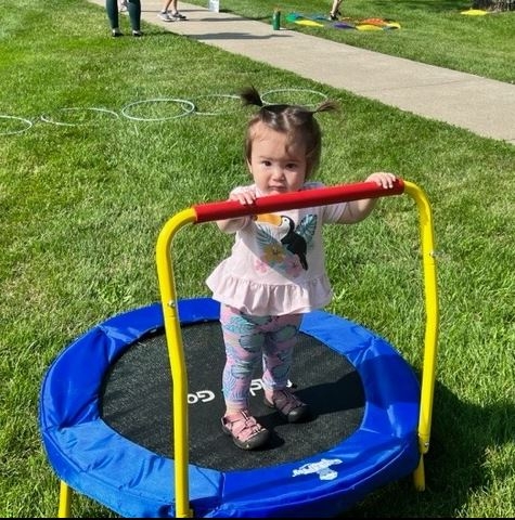 A toddler holding a railing and standing on a small trampoline on the South Lawn. 