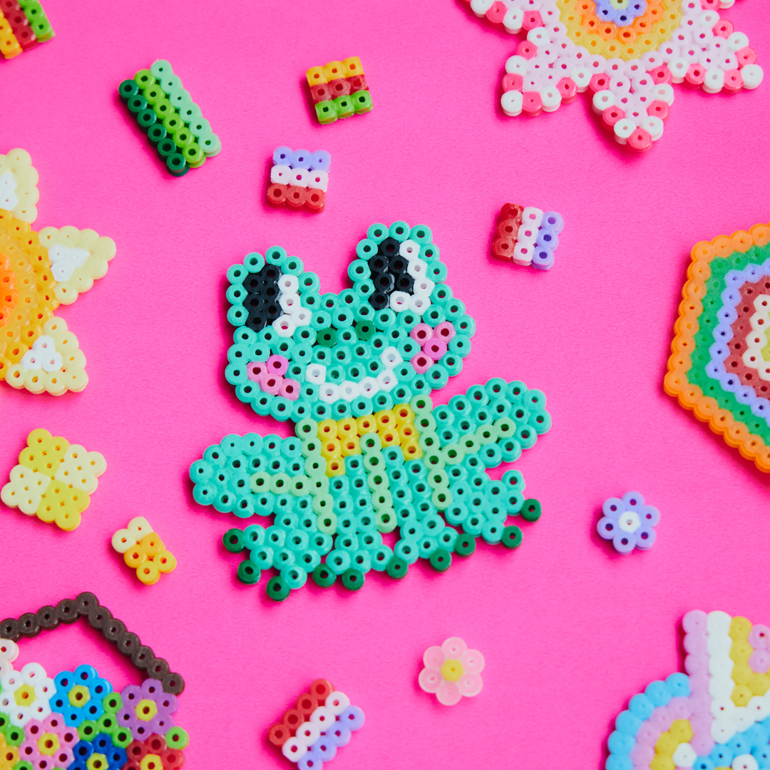 A Perler bead frog with a bright pink background and other Perler bead creations surrounding. 