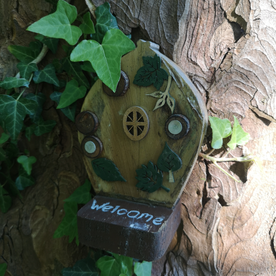A handmade fairy door against a tree, with greenery nearby. 