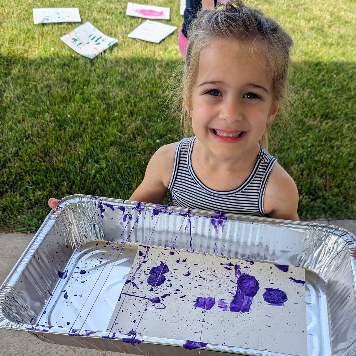 A girl holding up a tray covered in purple messy art, on the South Lawn of the Library. 