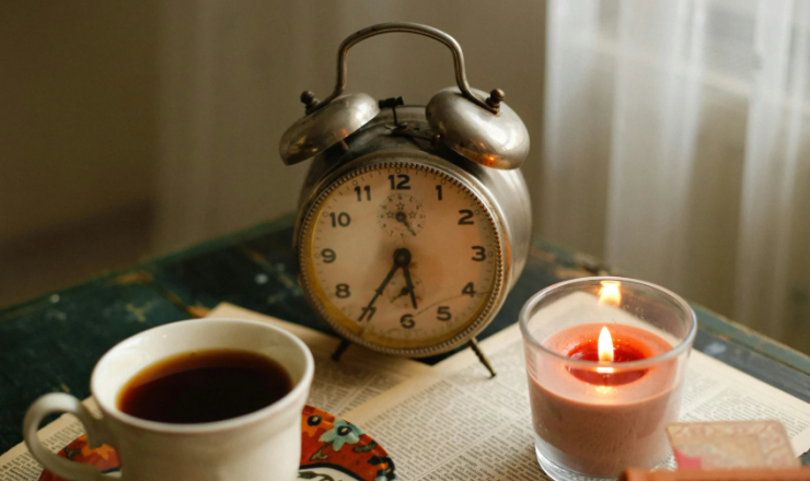 A clock on a table next to a cup of coffee and a lit candle. 